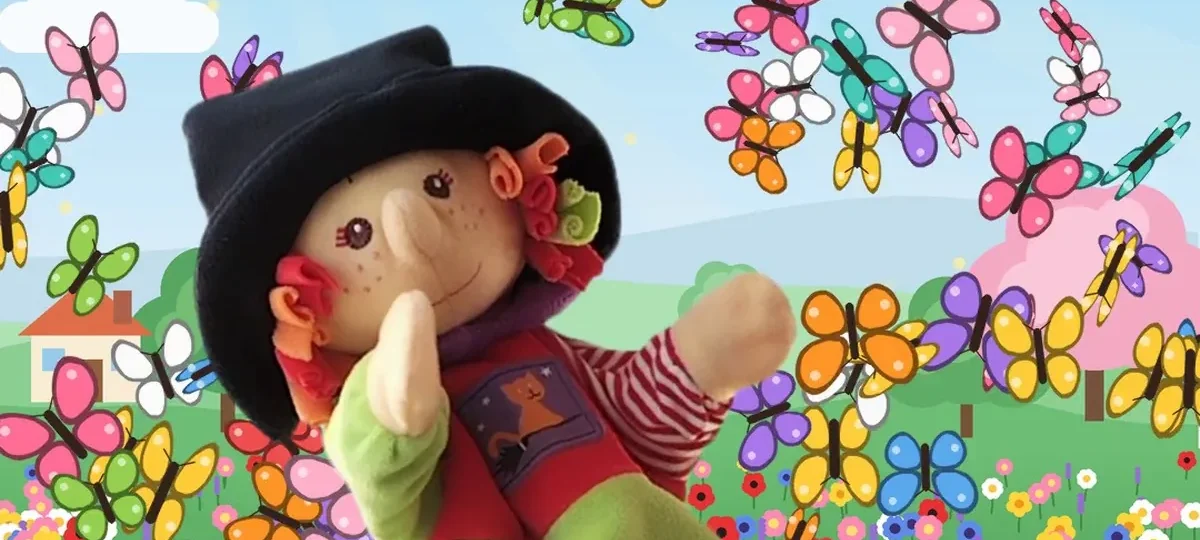 Marioneta de una brujita con sombrero negro, jersey rojo, falda verde y botas rojas, sentada en un tronco sobre un arroyo, rodeada de un torbellino de mariposas de colores y flores.