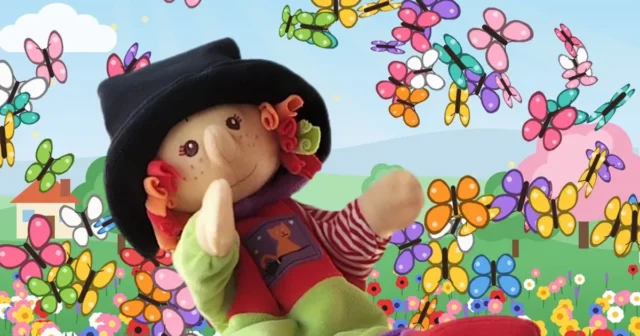 Marioneta de una brujita con sombrero negro, jersey rojo, falda verde y botas rojas, sentada en un tronco sobre un arroyo, rodeada de un torbellino de mariposas de colores y flores.